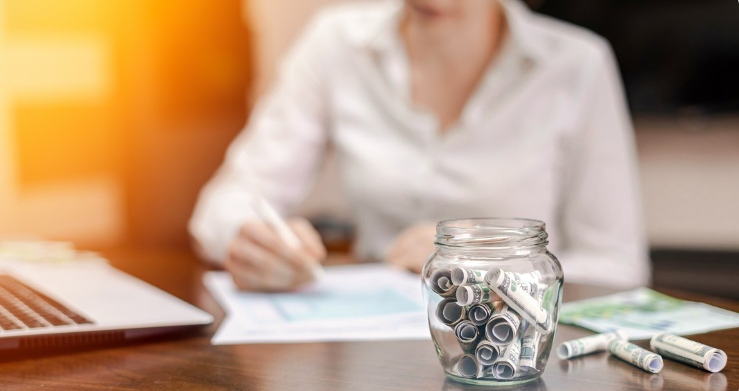 Close-up of a savings jar filled with cash on a wooden office desk, symbolizing wealth management and financial goals