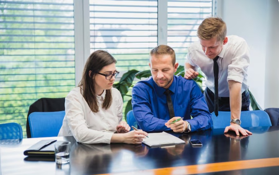 Three professionals are gathered around a black conference table reviewing a document on a clipboard