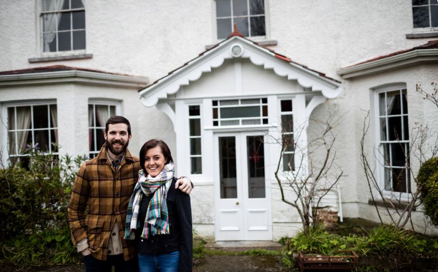 Young couple standing in front of a white house, smiling outside their home after purchasing residential property