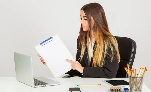 Professional woman reviewing documents on a clipboard while working on a laptop at an office desk