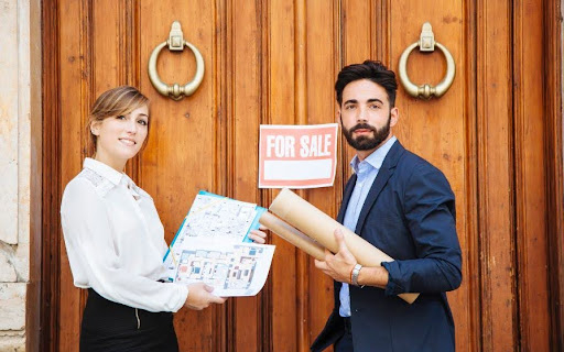 Real estate agents holding property plans in front of a house with a for sale sign