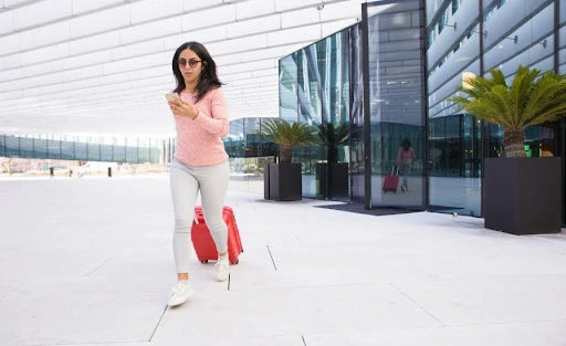 Woman with suitcase walking outside a modern office building while using her phone