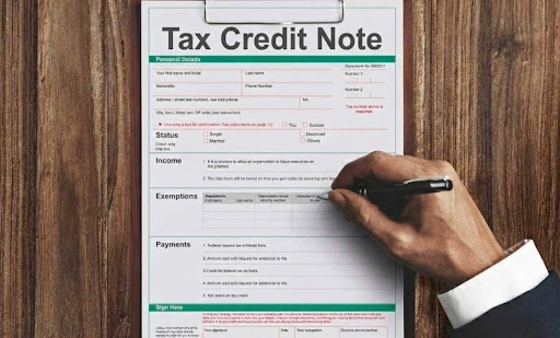 A person filling out a "Tax Credit Note" form on a clipboard, showing sections for income, exemptions, and payments.