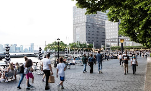 People walking along a waterfront promenade with cafes, boats, and modern city buildings in the background