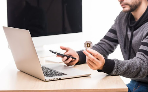 A close-up of a professional at a desk with a laptop and smartphone, holding a physical Bitcoin coin, representing UAE crypto regulation
