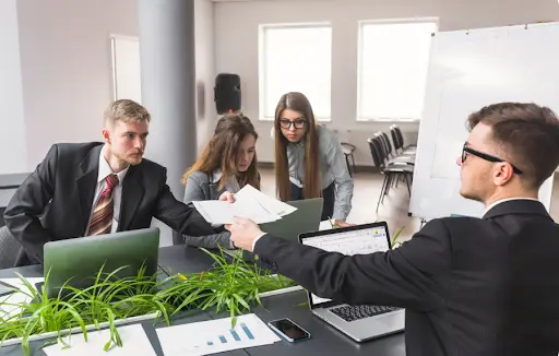 One team member is passing a stack of white papers to another across a desk
