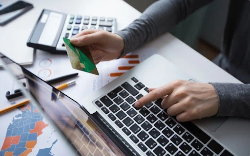 a person using a laptop while holding a green and gold credit card, with a financial calculator and data-driven charts visible on the desk