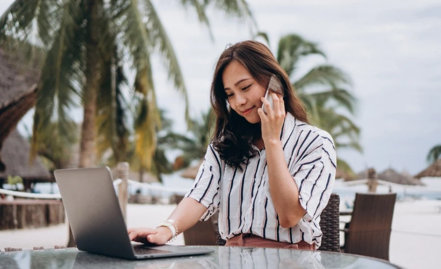 a woman sitting on a beach side and working on a laptop