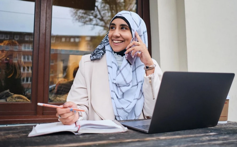 Young smiling arabic woman in hijab happily talking on smartphone while working on laptop